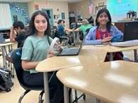  Two female students, sitting at a table with their laptops, learning how to code.